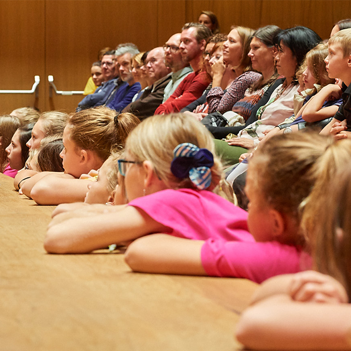 Kinder und Erwachsene im Publikum, auf eine Bühne blickend