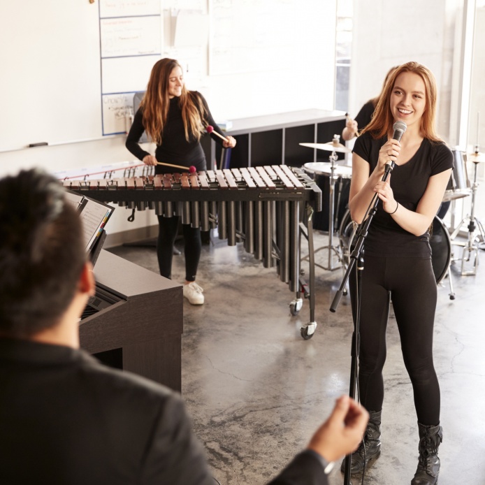 eine Band beim Proben, bestehend aus Mädchen am Mikrophon, Mädchen auf einer Marima spielend, Drumset im Hintergrund und Dirigent