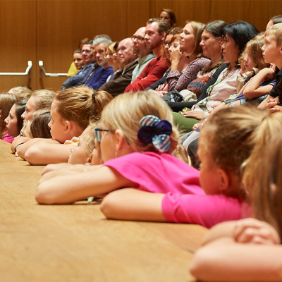 Kinder und Erwachsene im Publikum, auf eine Bühne blickend