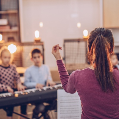 Mädchen in Rückenansicht dirigierend vor einer verschwommenen Gruppe musizierender Kinder, zwei davon am Keyboard