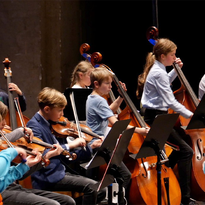 Junge Orchestermusiker:innen auf ihren Streichinstrumenten inmitten eines Orchesterauftritts