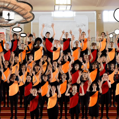 Kinderchor in orange-schwarz-roten Kostümen mit erhobenen Armen auf einer Treppe stehend